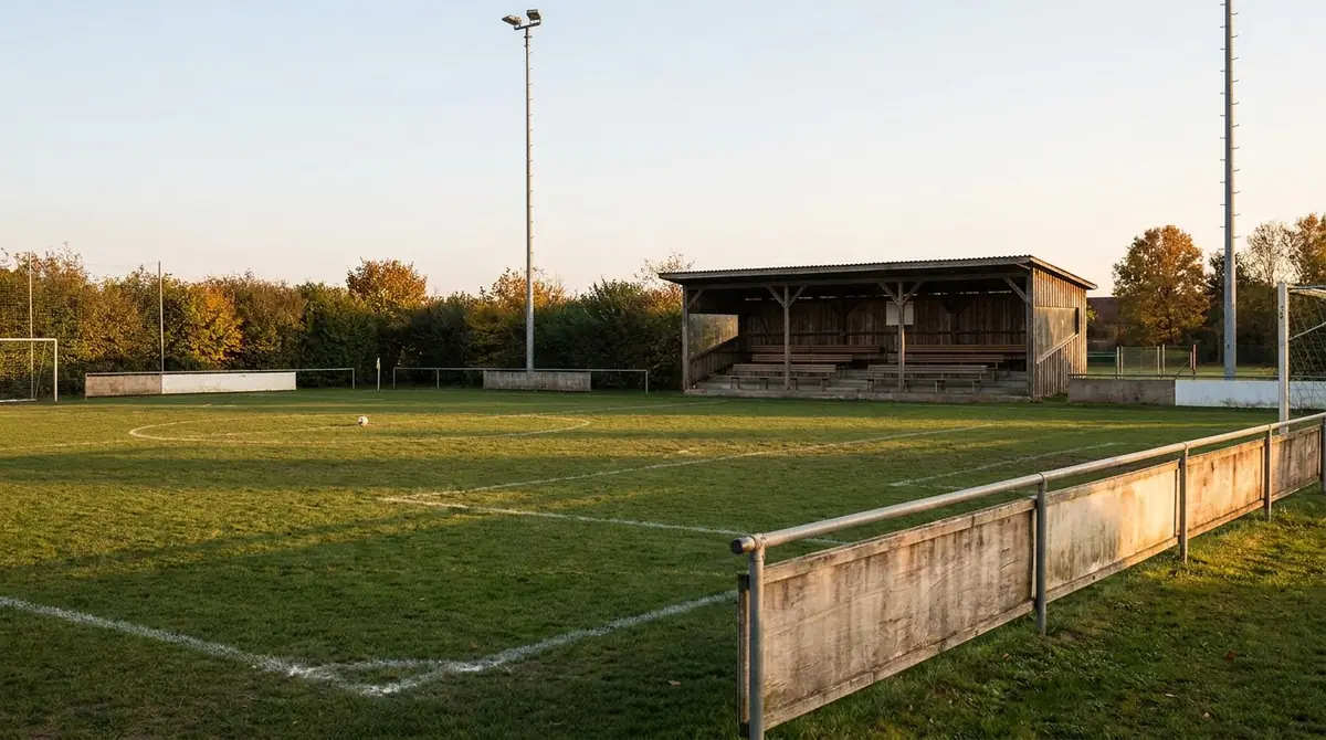 Amateur-Fußballplatz mit Rasen, Flutlichtmast, überdachter Holztribüne und Ball auf dem Anstoßpunkt